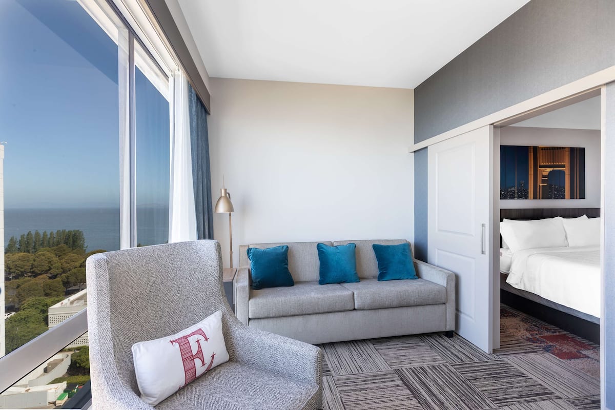 Une chambre d’hôtel moderne avec une grande fenêtre, un canapé gris agrémenté de coussins bleus, un lit blanc et une moquette à motifs.