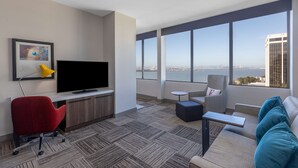 Une chambre d’hôtel moderne dotée d’une grande fenêtre donnant sur la ville, d’un téléviseur à écran plat, d’un fauteuil de bureau rouge, d’un canapé gris et d’une petite table ronde.