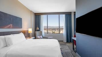A hotel room with a large bed, a desk, a chair, a TV, and a window with a city view.