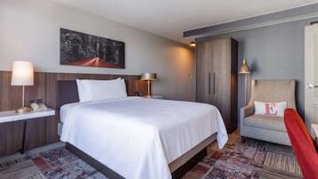A hotel room with a large bed, a chair, a desk with a lamp, and a painting on the wall.