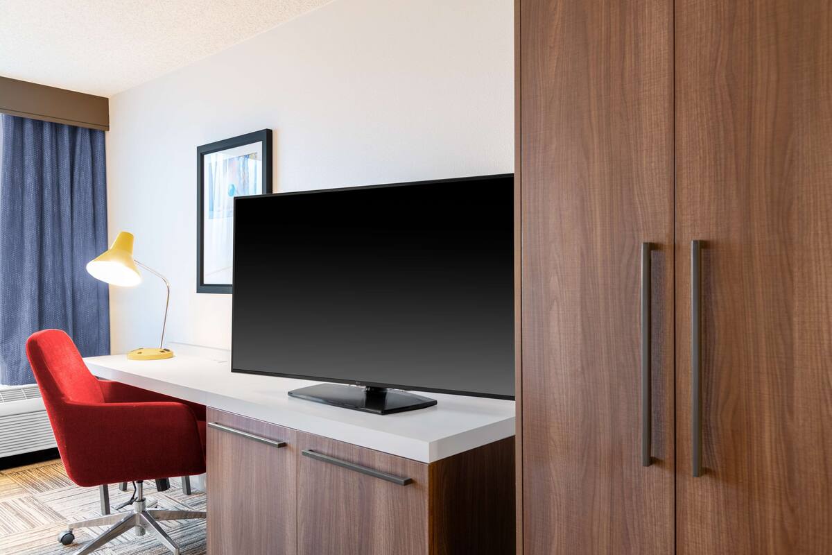 Een hotelkamer met een bureau, een rode stoel, een flatscreen televisie, een lamp en een kledingkast.