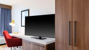 A hotel room with a desk, a red chair, a flat-screen TV, a lamp, and a wardrobe.