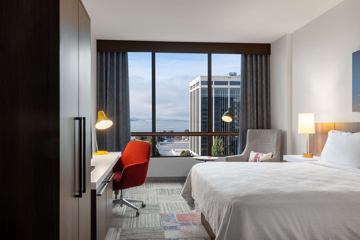 Une chambre d’hôtel avec un grand lit, un bureau, une chaise et une vue sur le paysage urbain, grâce à de grandes fenêtres.