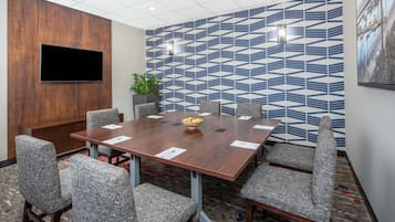 Uma sala de conferências com uma grande mesa de madeira, cadeiras estofadas cinzentas, uma TV de tela plana fixada em um painel de madeira e uma parede estampada.