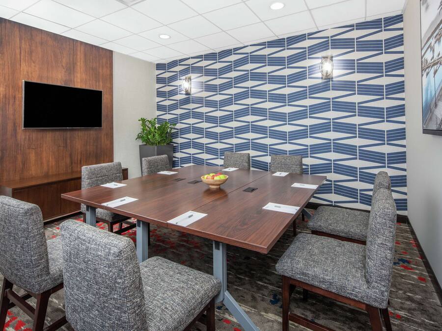 A conference room with a large wooden table, grey upholstered chairs, a flat-screen TV mounted on a wooden panel, and a patterned wall.