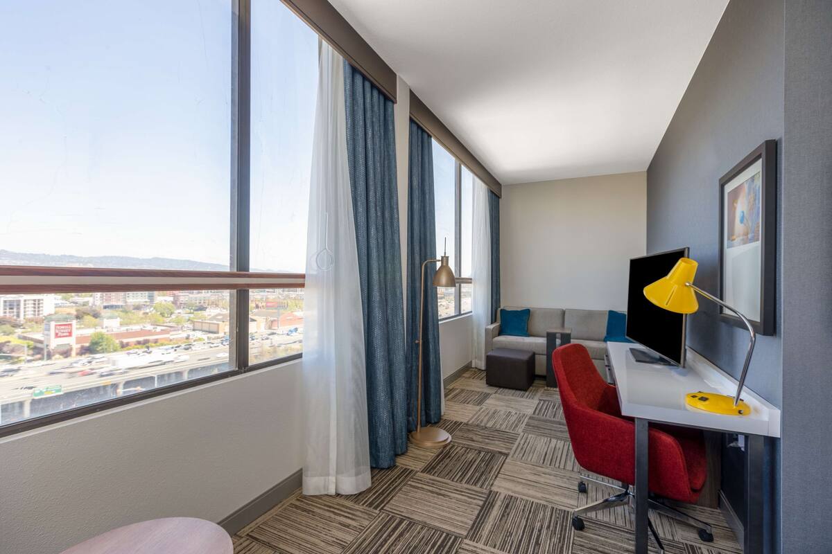 Une chambre d’hôtel moderne avec une grande fenêtre, un bureau doté d’une lampe jaune, une chaise rouge et un canapé avec des coussins bleus.