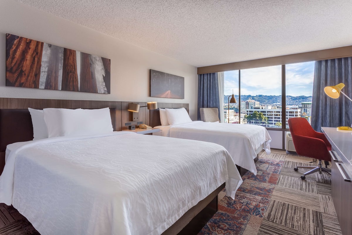 Une chambre d’hôtel avec deux lits, un bureau, une chaise, une lampe et une grande fenêtre donnant sur la ville.