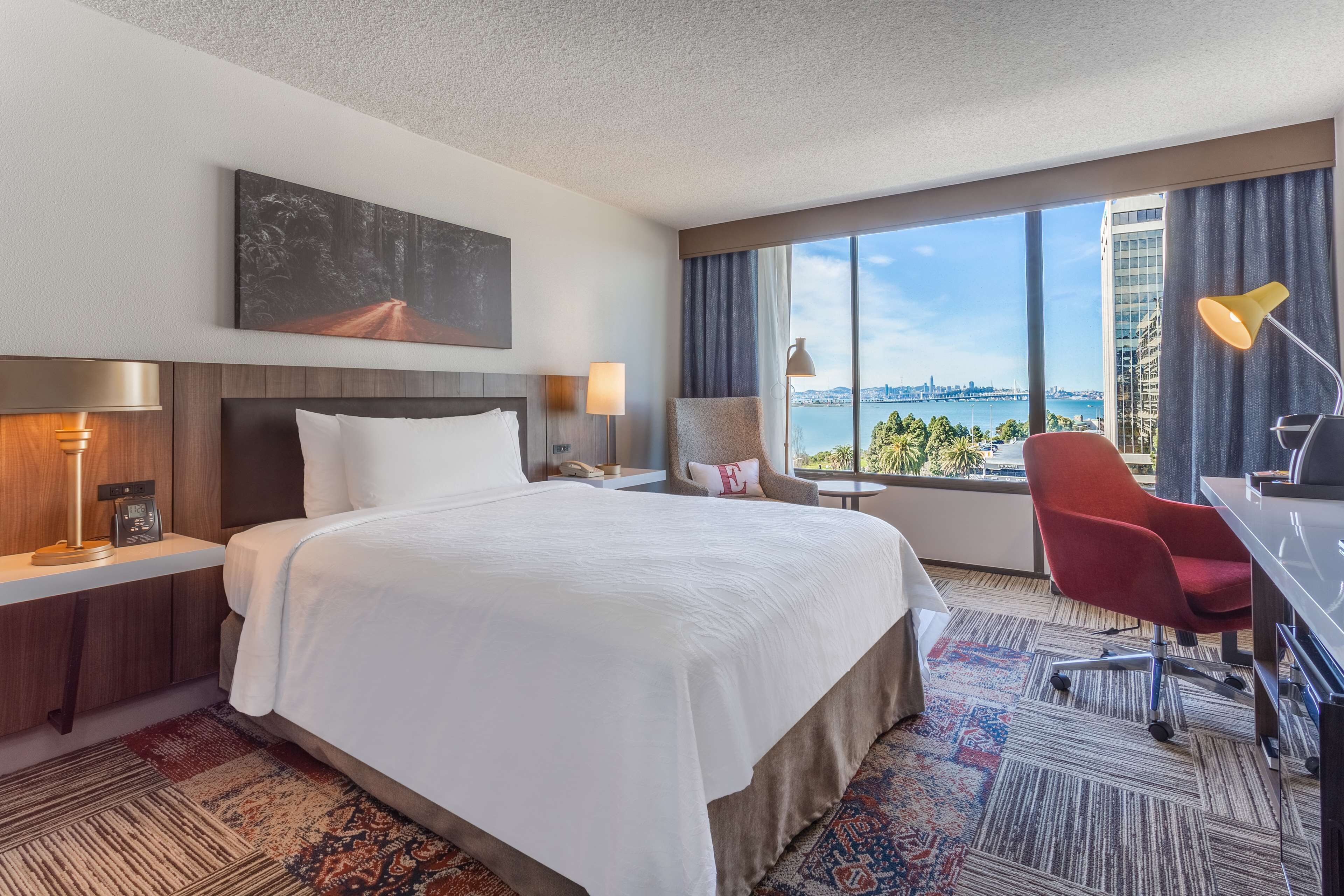 A hotel room with a large bed, a desk with a chair, a view of a cityscape, and a large window with curtains.
