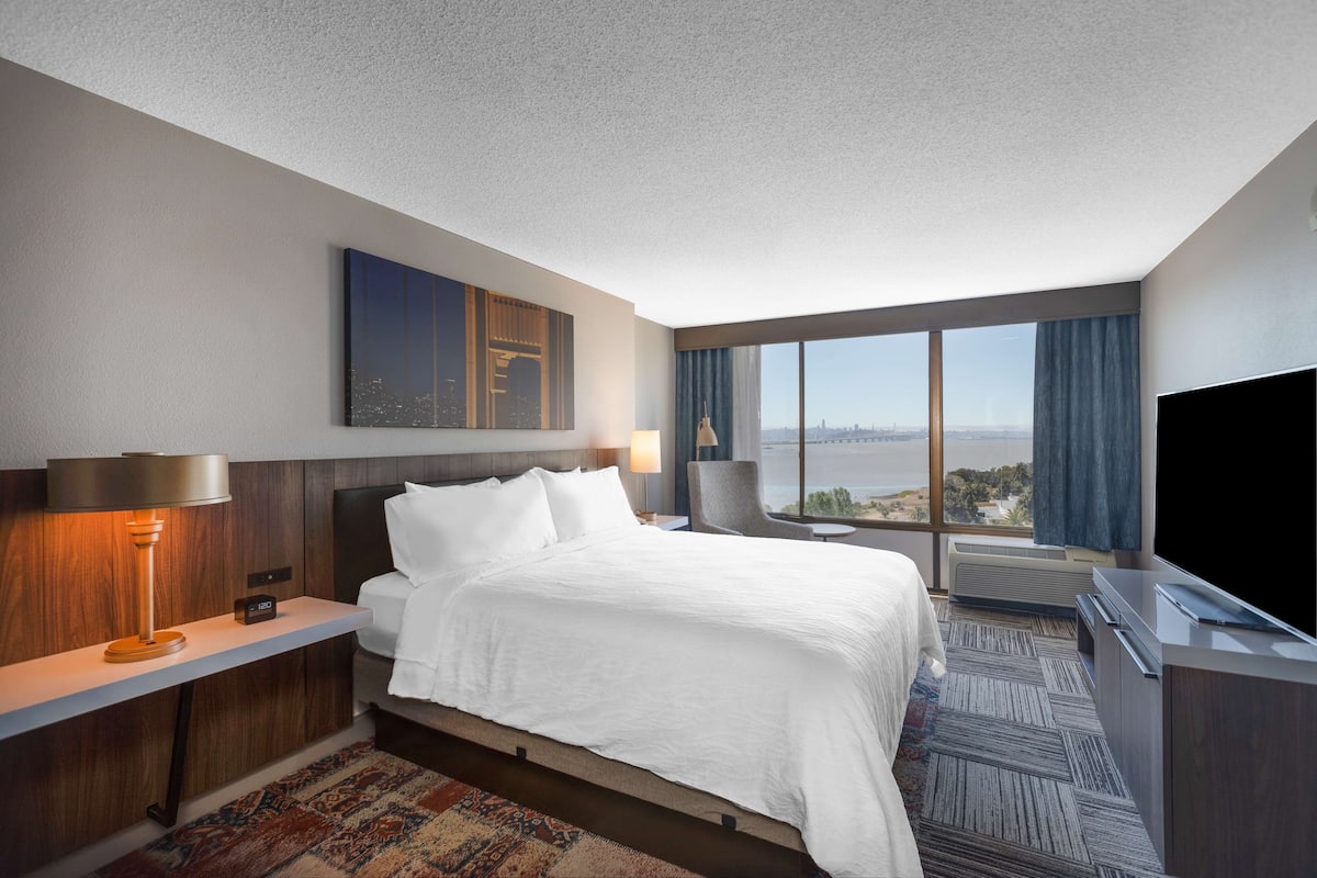 Une chambre d’hôtel dotée d’un grand lit, d’un bureau avec une lampe, d’un téléviseur à écran plat et d’une grande fenêtre donnant sur la ville.