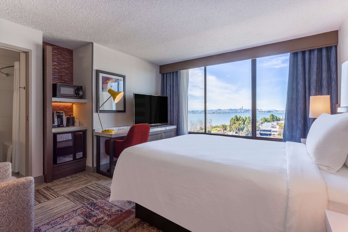 Une chambre d’hôtel avec un grand lit, un bureau avec un ordinateur, une télévision et une vue sur le paysage urbain grâce à de grandes fenêtres.