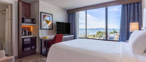 A hotel room with a large bed, a desk with a computer, a TV, and a view of a cityscape through large windows.