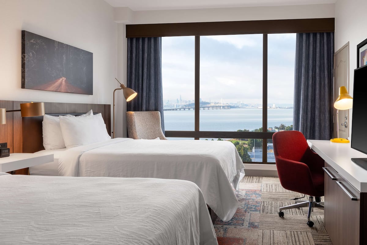 Une chambre d’hôtel avec deux lits, un bureau, une chaise, une télévision et une grande fenêtre donnant sur la ville.