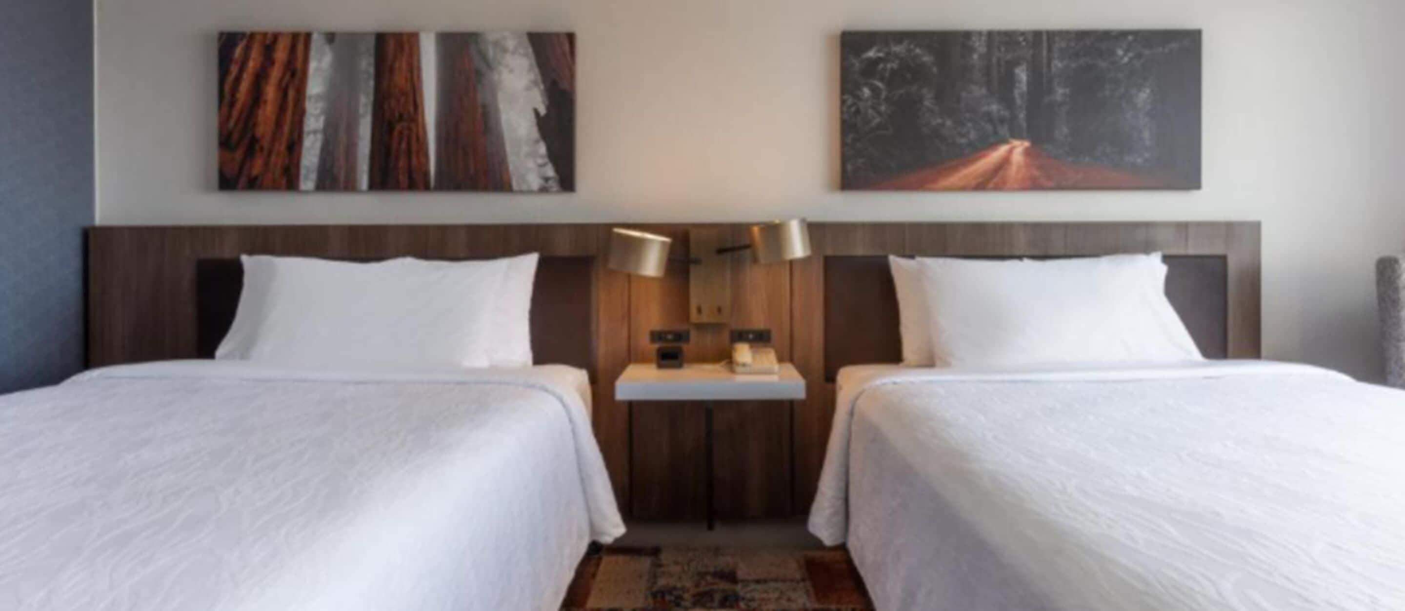 A hotel room with two beds, a nightstand, and two wall-mounted lamps.