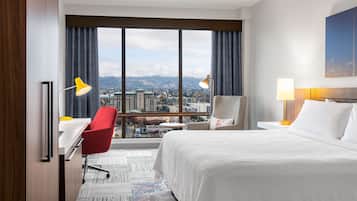 A hotel room with a large window, a bed, a desk with a chair, and a nightstand with a lamp.
