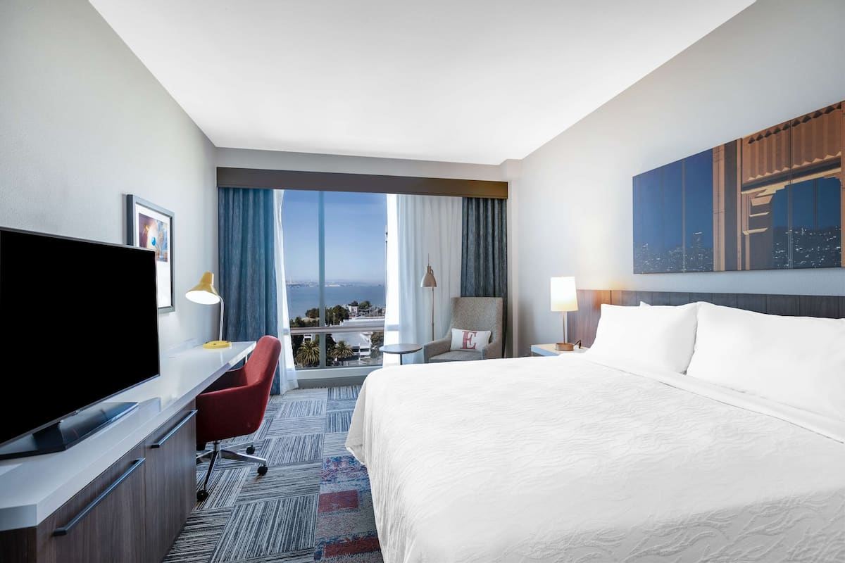 Une chambre d’hôtel avec un grand lit, un bureau doté d’un téléviseur à écran plat, une chaise, une table de chevet avec une lampe, et une vue sur le paysage urbain par la fenêtre.