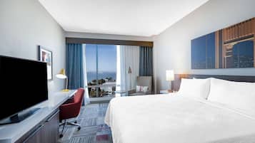 A hotel room with a large bed, a desk with a flat-screen TV, a chair, a nightstand with a lamp, and a cityscape view through the window.