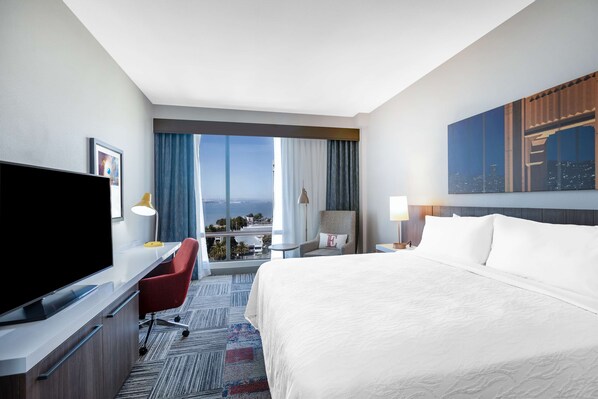 Habitación de hotel con una cama grande, un escritorio, un televisor de pantalla plana, una silla, una mesita de noche con lámpara y vistas a la ciudad a través de la ventana.