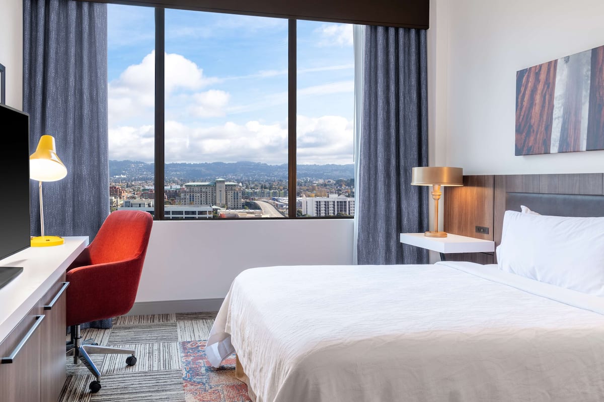 Une chambre d’hôtel avec une grande fenêtre, un lit, un bureau avec une chaise et une lampe.