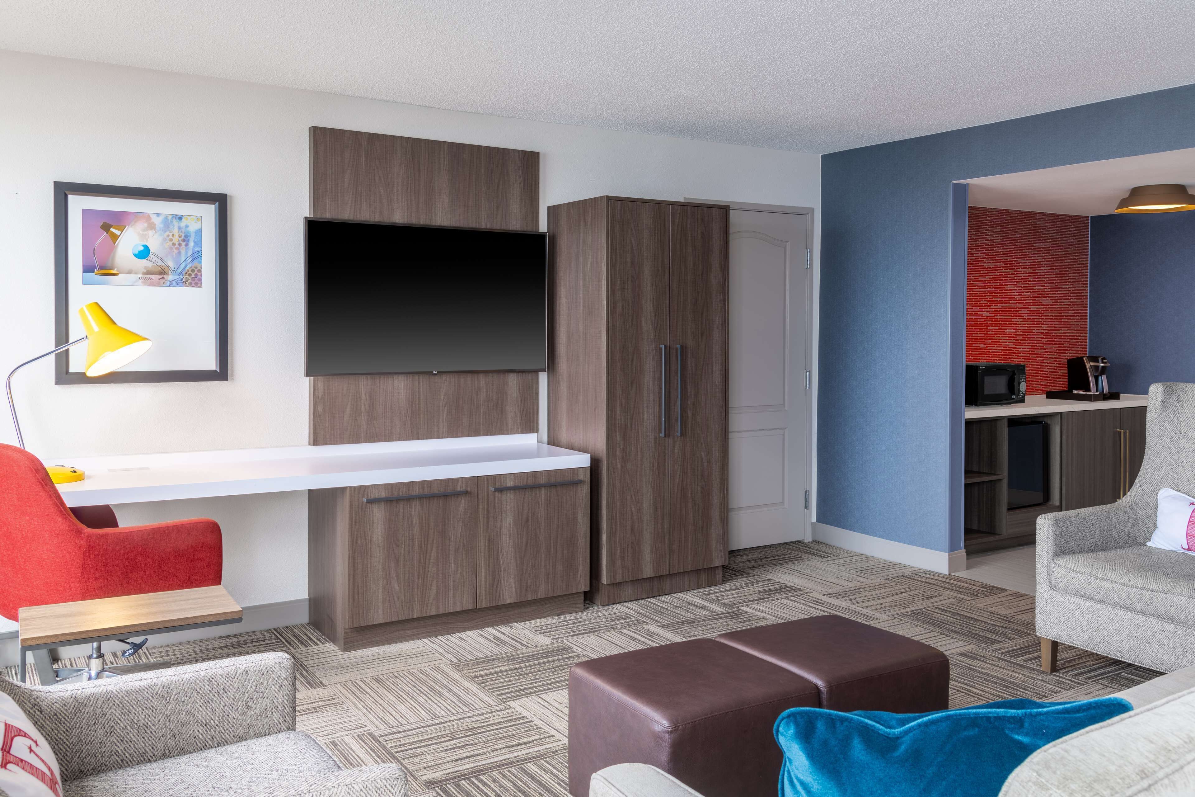 Una habitación moderna con televisor de pantalla plana, armarios de madera, kitchenette y un área de descanso con una silla roja y un sofá.