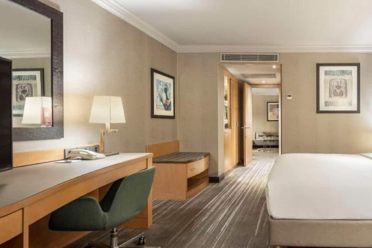 A hotel room with a bed, a desk with a chair, a mirror, and a bedside table with a lamp.