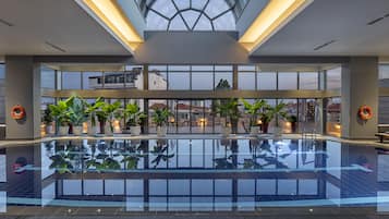 Una amplia piscina interior con paredes de vidrio transparente, plantas en macetas y sillones de descanso.