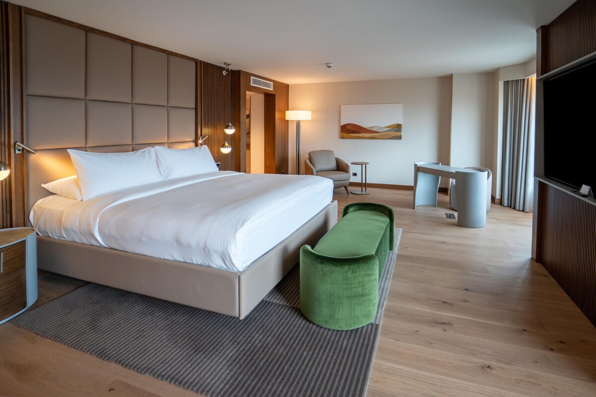 Una camera d'albergo moderna con un letto grande, un poggiapiedi verde, una scrivania e una televisione.