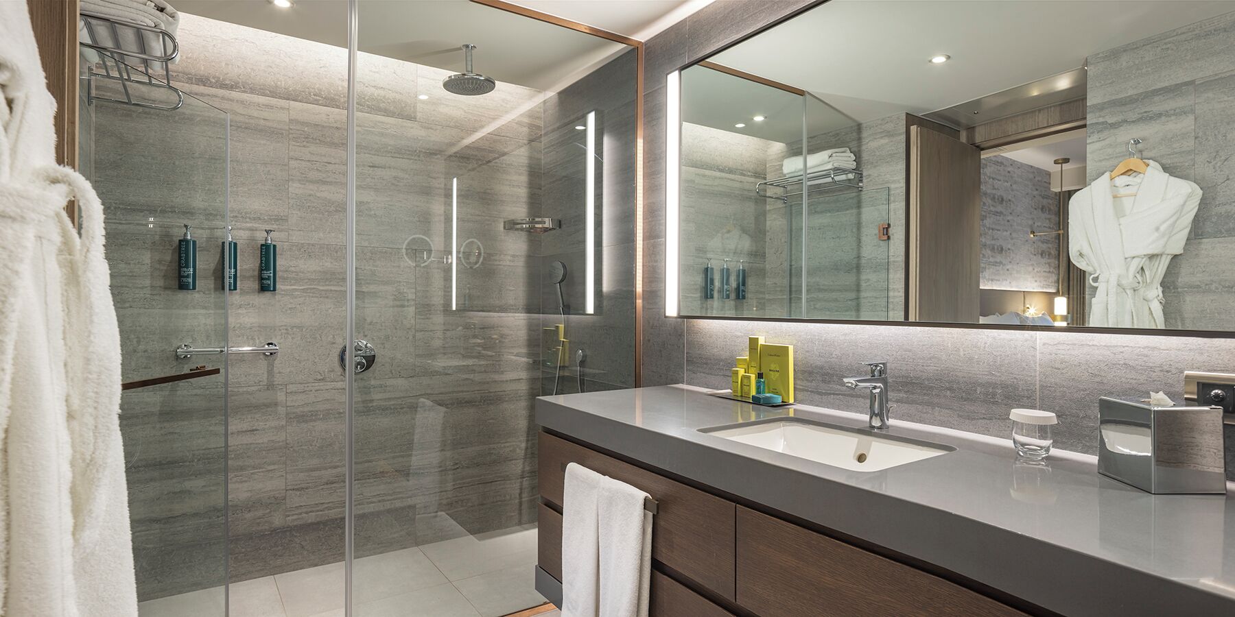 Un baño moderno con ducha de vidrio, un espejo grande y un tocador con lavamanos y artículos de aseo.
