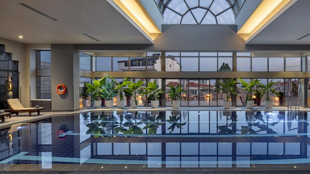 A large indoor swimming pool with clear glass walls, potted plants, and lounge chairs. - Ankara HiltonSA (Ankara)