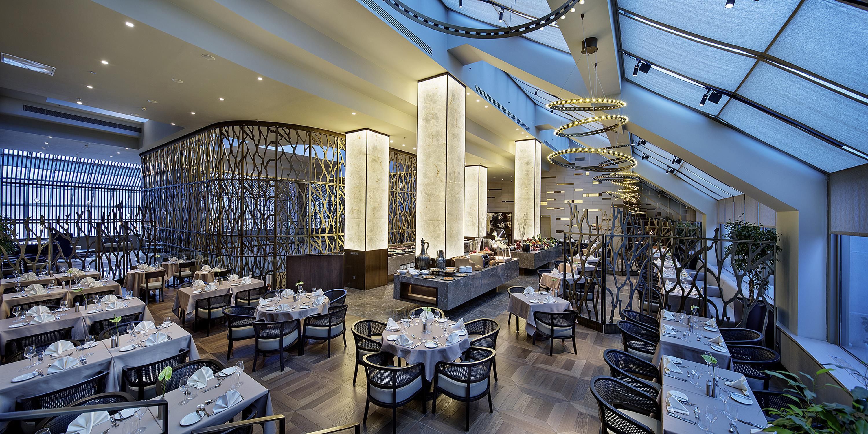 A modern restaurant with multiple dining areas, high ceilings, and contemporary lighting fixtures.