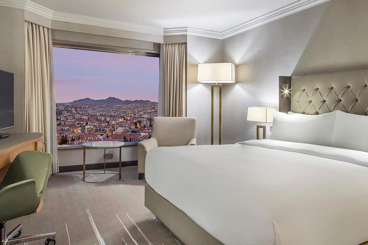 A hotel room with a large bed, a desk with a chair, a small table, and a view of a cityscape.