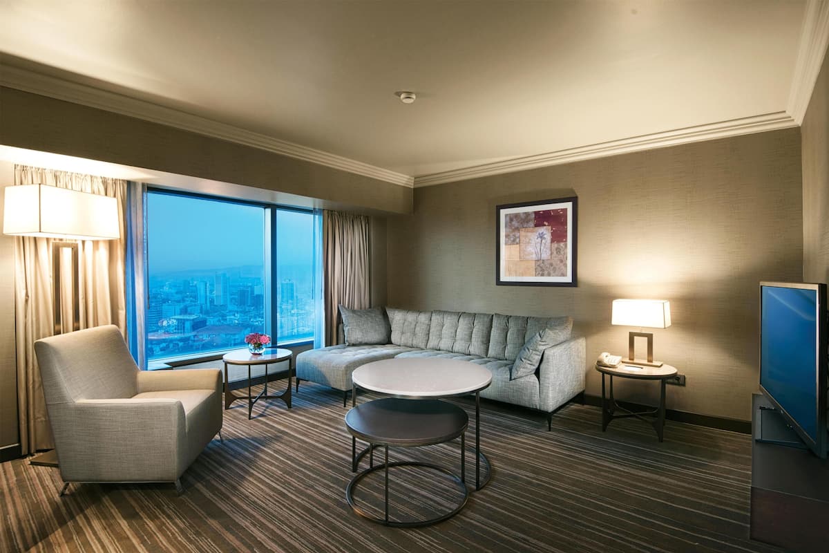 A hotel room with a large window, a sofa, a round table, a chair, a TV, and a lamp.