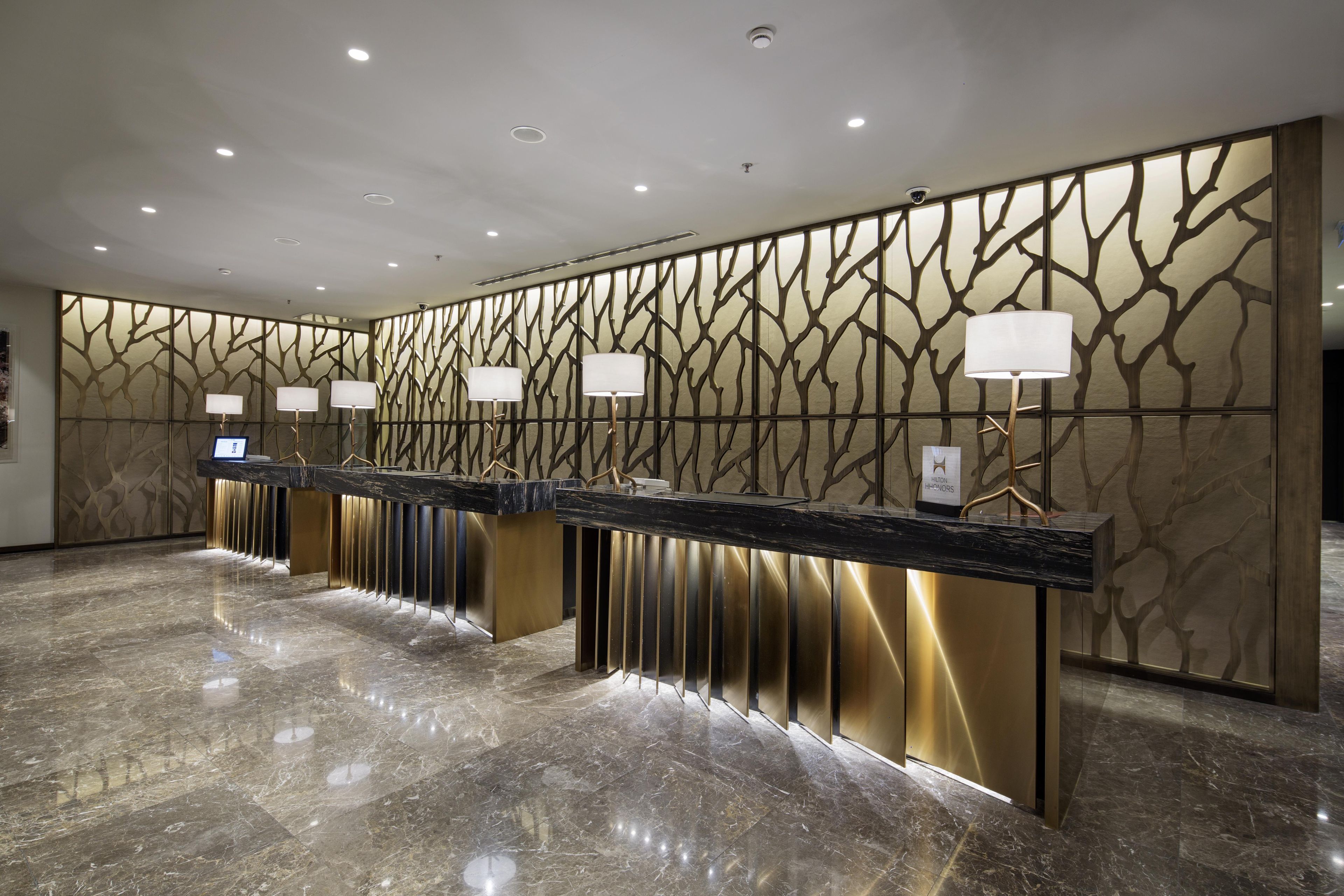 Een hotel receptie met een marmeren vloer, een receptiebalie met een digitaal scherm en een decoratieve muur met een boommotief.