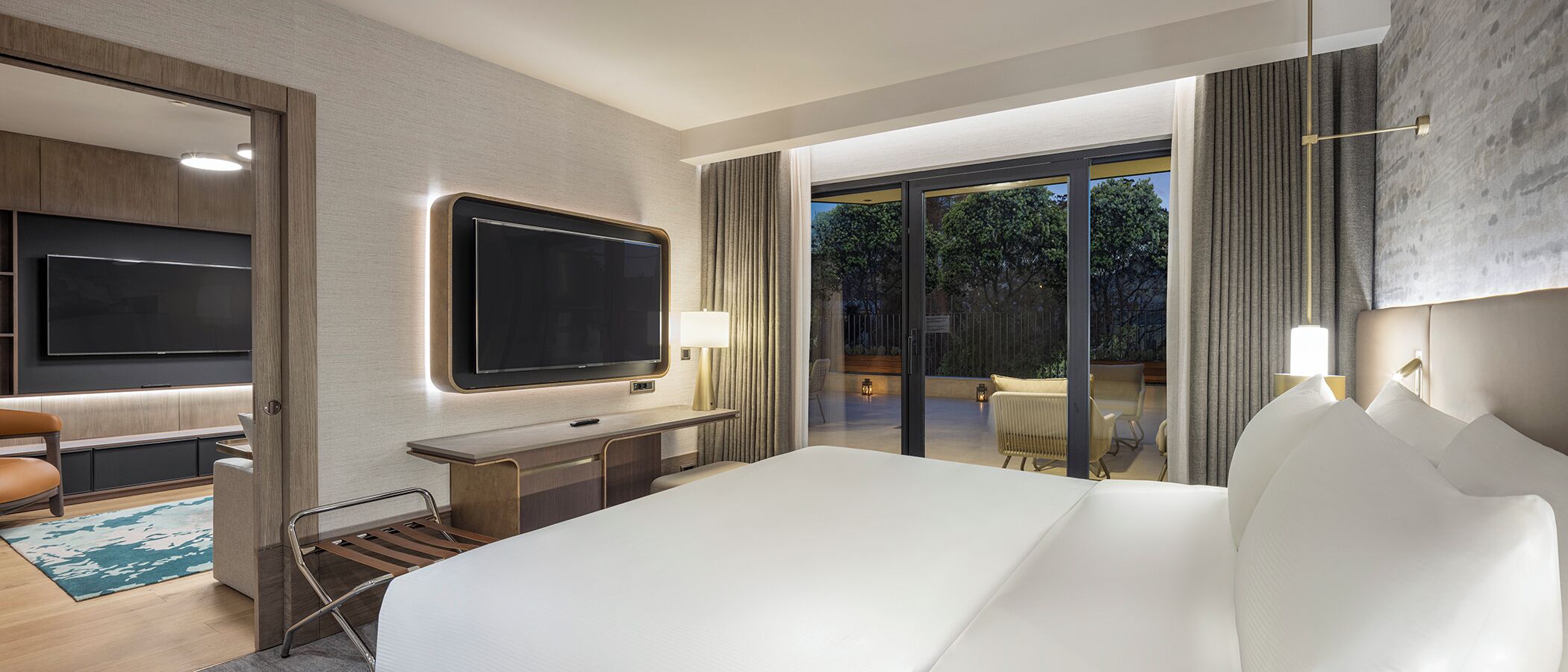 A hotel room with a large bed, a desk with a flat-screen TV, a seating area with chairs, and a view of the outdoors.