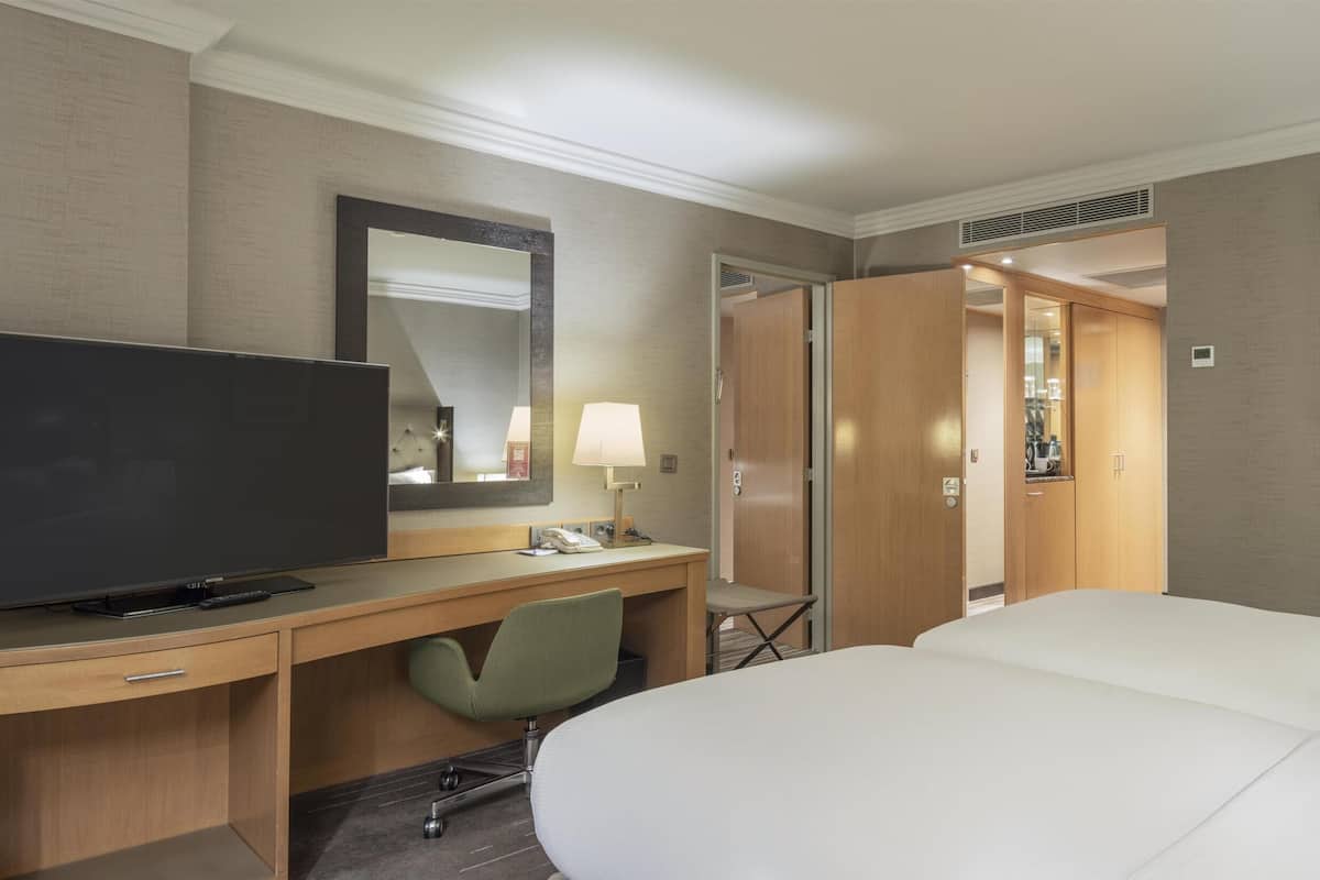 A hotel room with a bed, a desk with a TV, a chair, a lamp, and a framed picture on the wall.