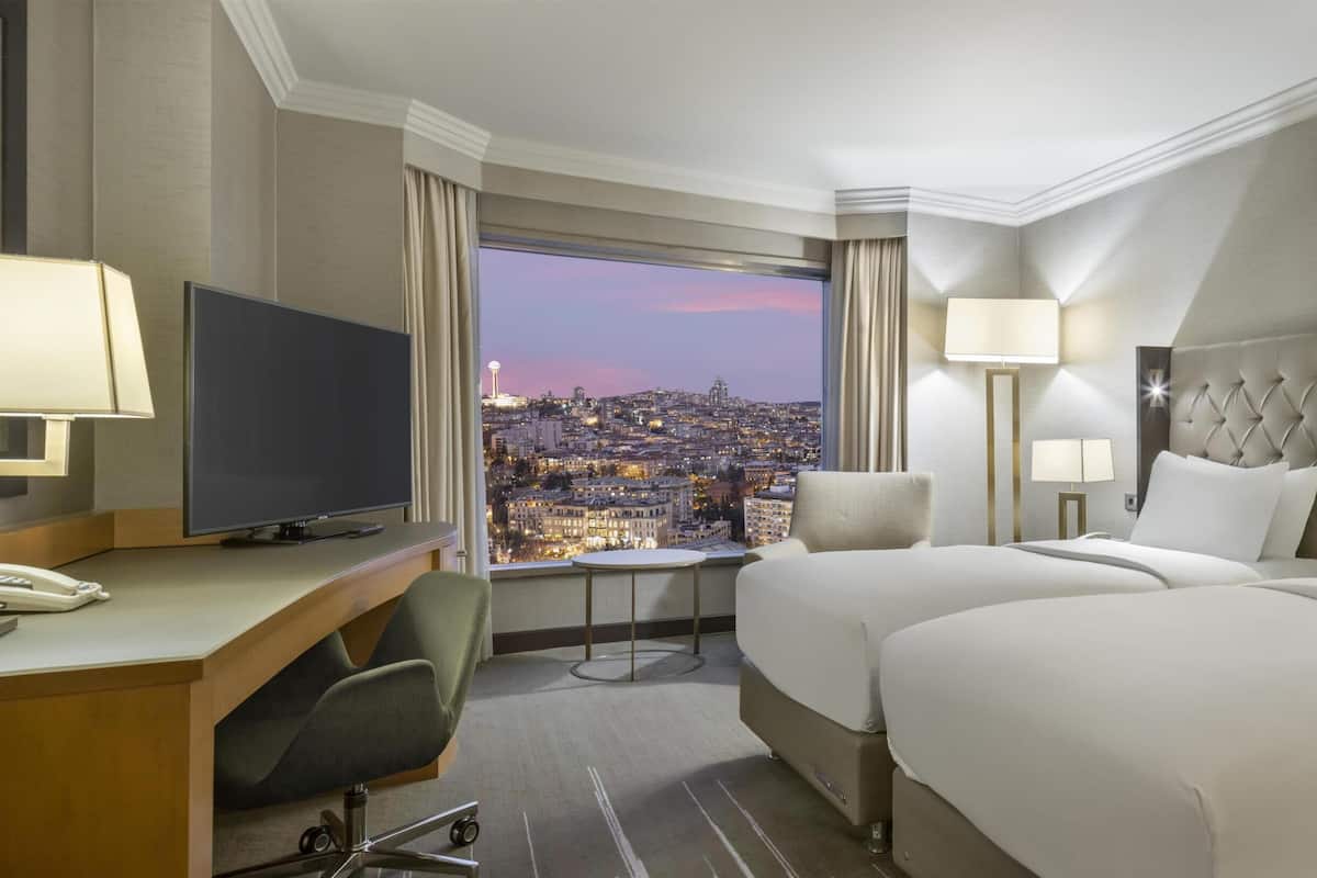 A hotel room with two beds, a desk with a TV, a chair, a small table, and a view of a cityscape at dusk.