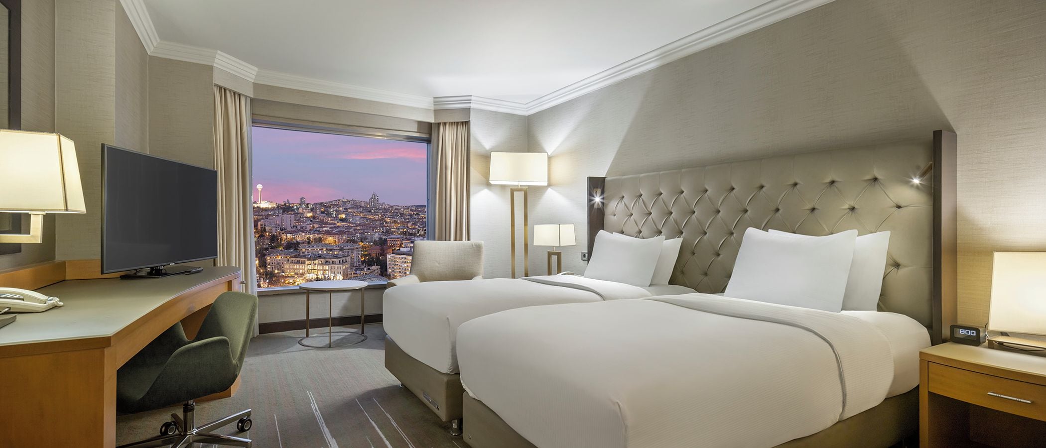 A hotel room with two beds, a desk, a chair, a television, and a view of a cityscape.