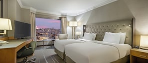 Habitación de hotel con dos camas, un escritorio, una silla, un televisor y vistas a un paisaje urbano.