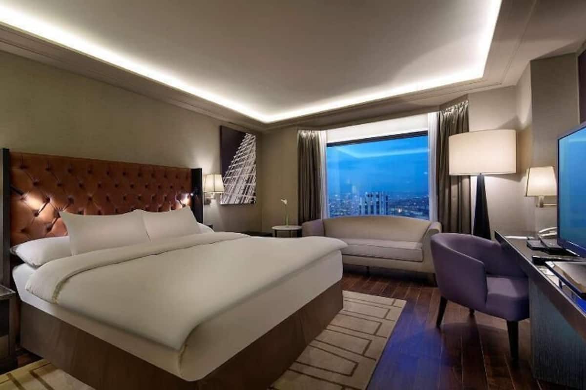 A hotel room with a large bed, a desk with a TV, a chair, and a view of the city.