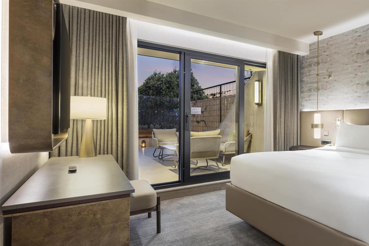 A hotel room with a large bed, a desk, a chair, a glass door leading to an outdoor area, and a modern light fixture.