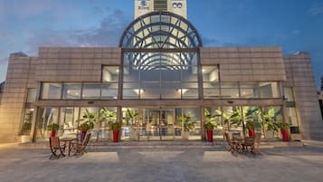 A hotel with a modern glass facade, a prominent logo at the top, and outdoor seating with potted plants.