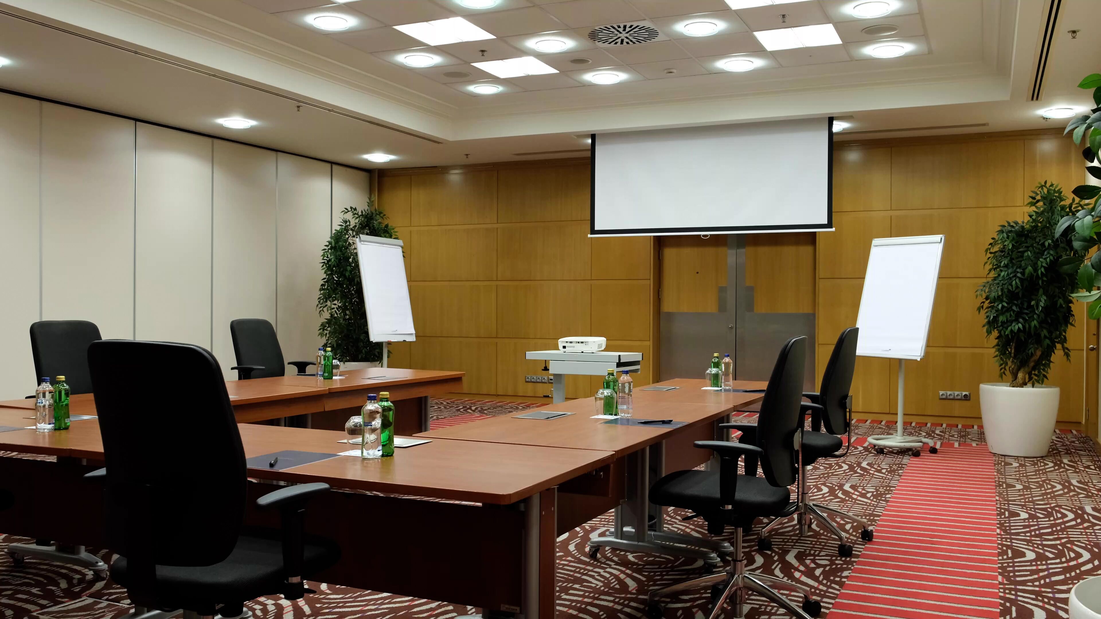 A conference room with a long table, multiple chairs, a projector screen, whiteboards, and potted plants.