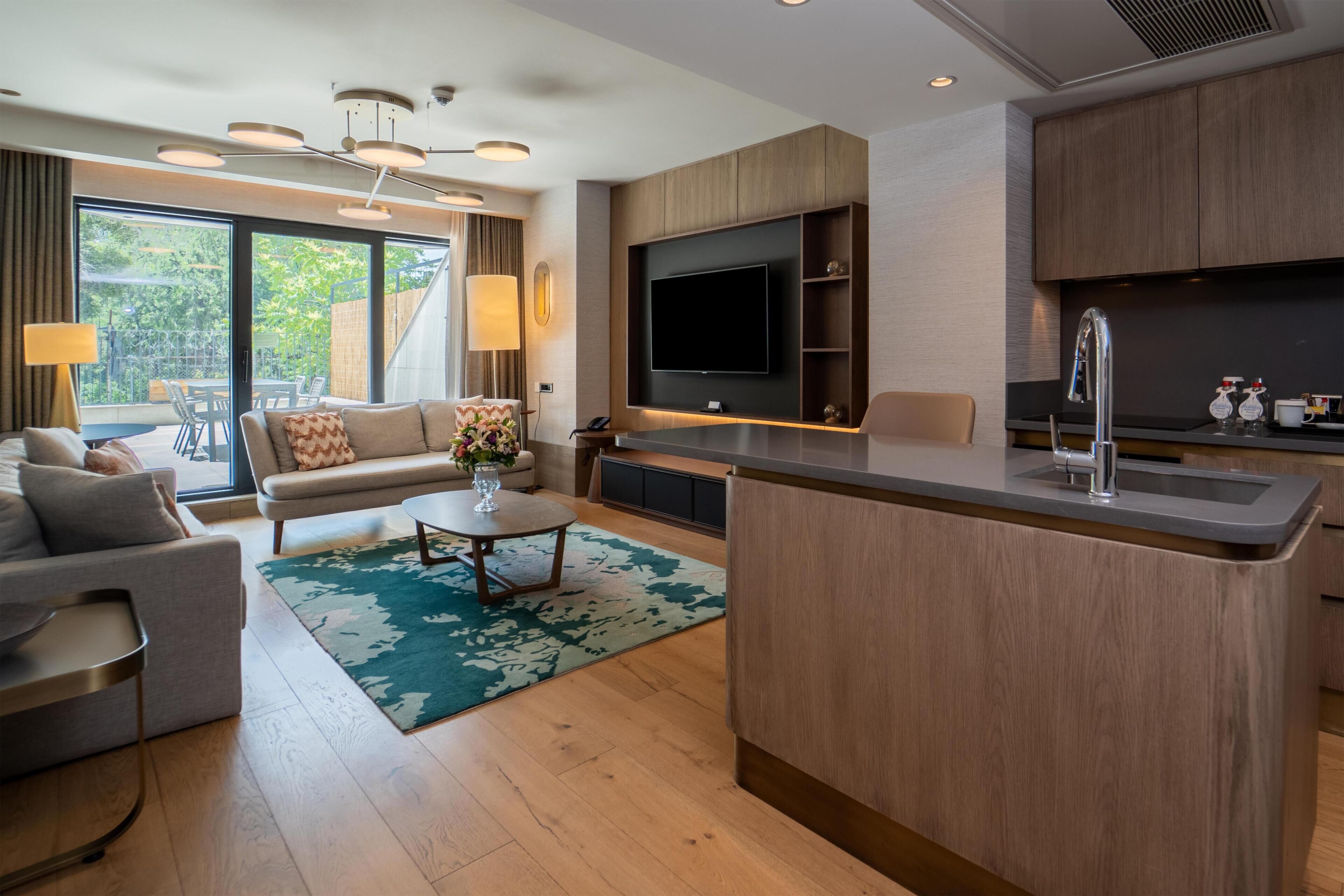A modern hotel room with a living area, kitchenette, and a large window offering a view of greenery.