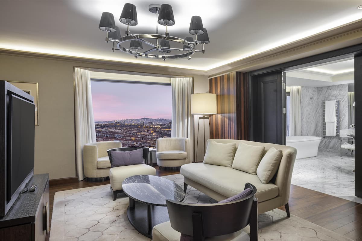 A modern hotel room with a large window, a sofa, a coffee table, a TV, and a chandelier.