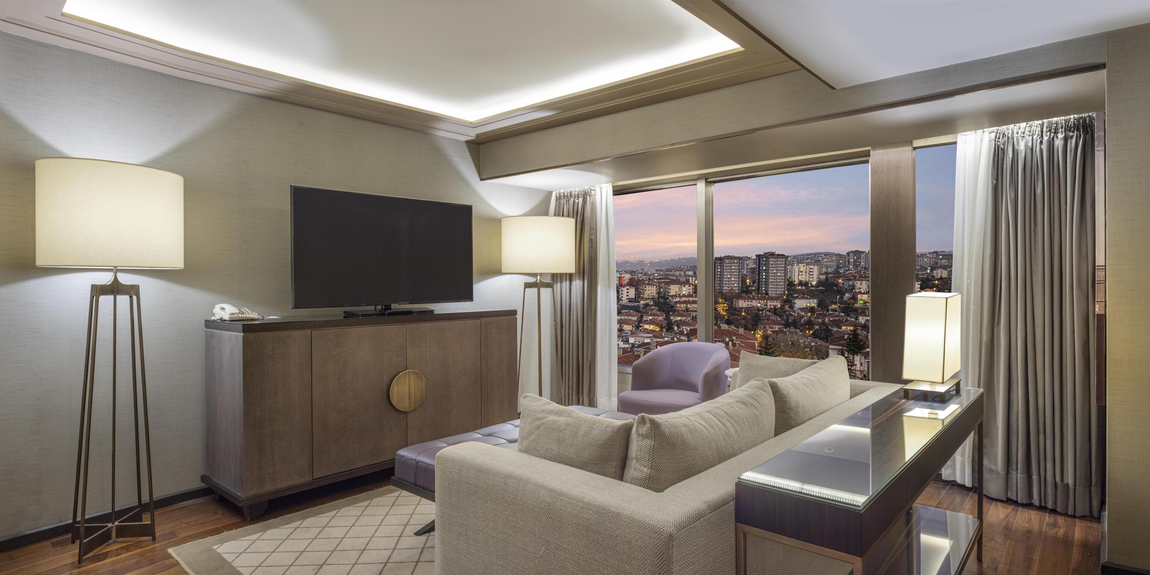 A modern hotel room with a large window, a flat-screen TV, a sofa, a desk, and two floor lamps.