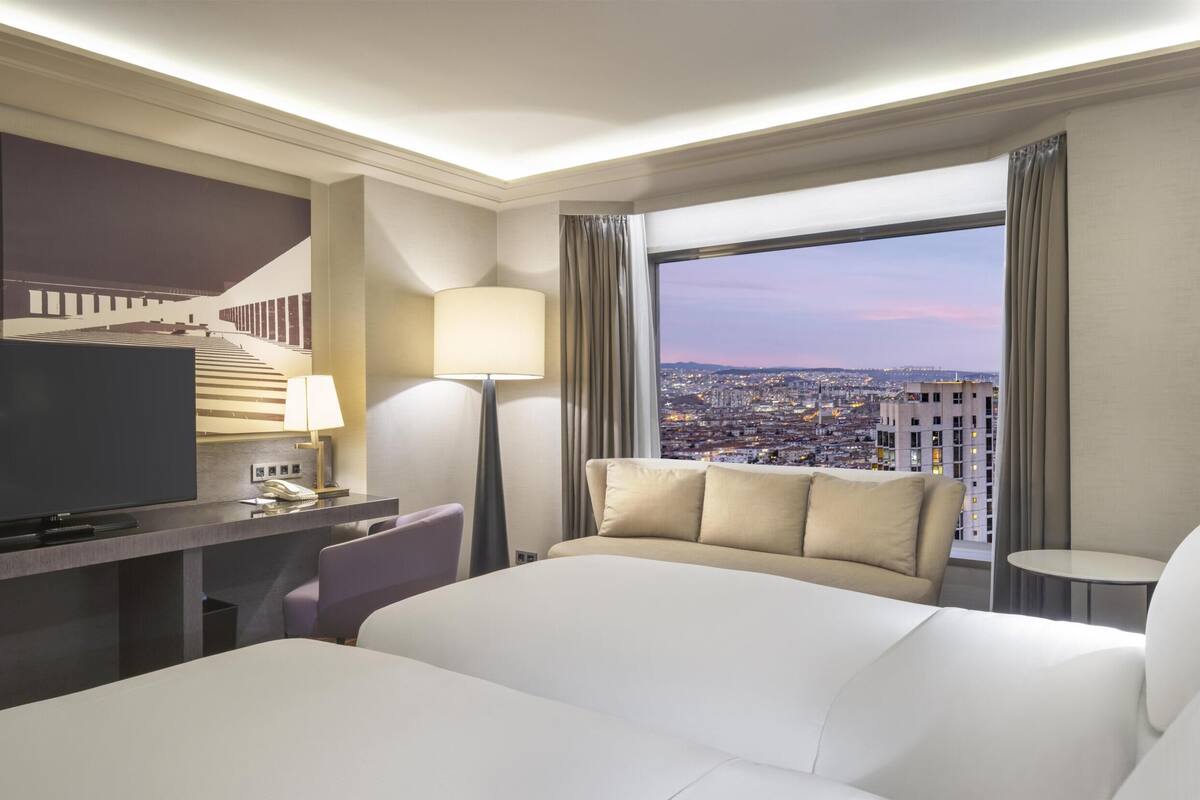 A hotel room with a bed, a TV, a desk, a chair, a lamp, and a view of a cityscape.