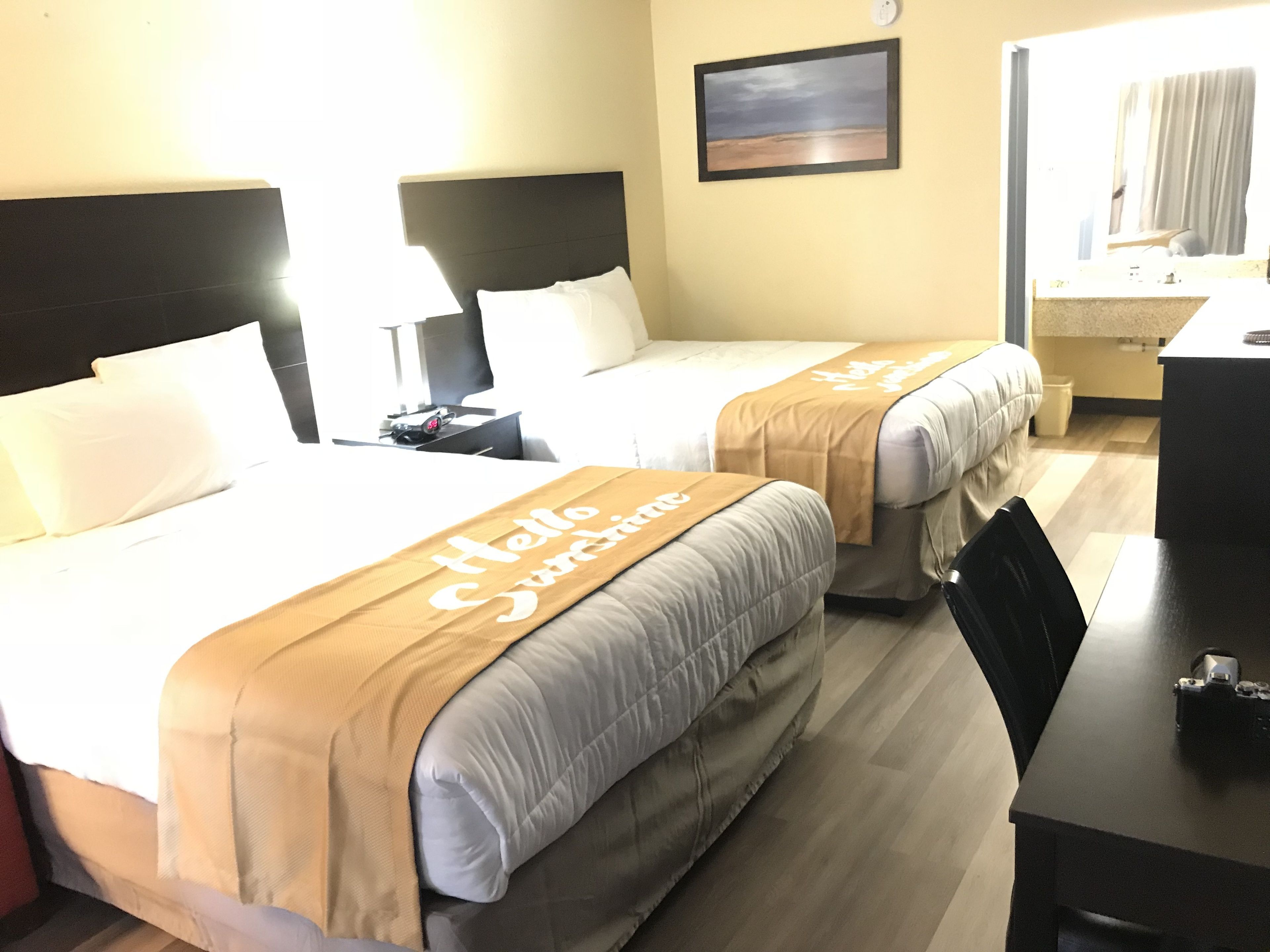 A hotel room with two beds, each with a 'Hello Sunshine' bedspread, a nightstand with a phone, a framed picture on the wall, and a desk with a camera.