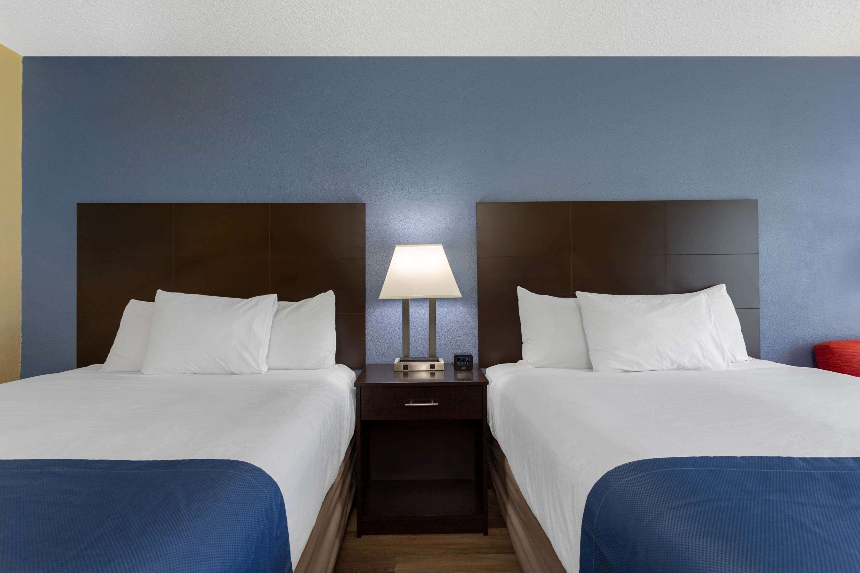 A hotel room with two beds, a nightstand with a lamp, and a red chair.
