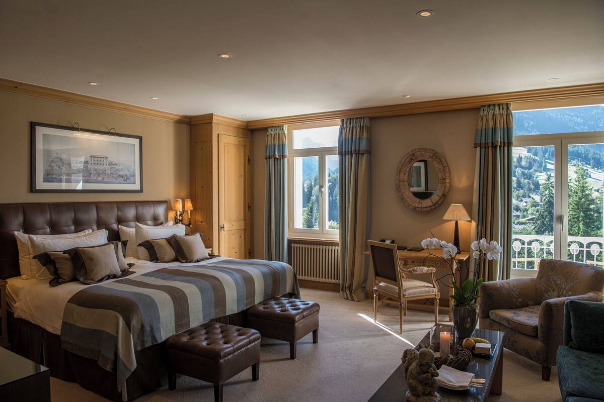 A spacious bedroom with a large bed, a sitting area with a chair and a table, and a view of the mountains through the windows.