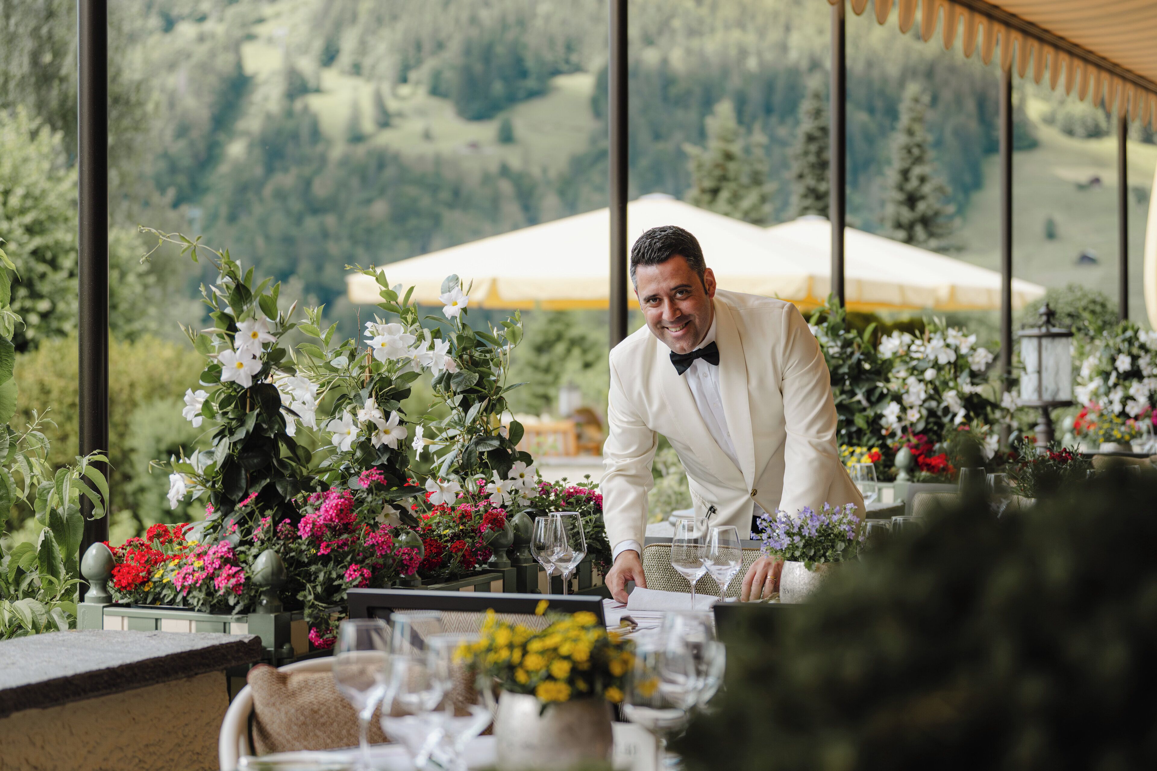 Een man in een wit pak die een tafel inricht met wijnglazen en bloemen, met uitzicht op een schilderachtige omgeving buiten, met een tent op de achtergrond.