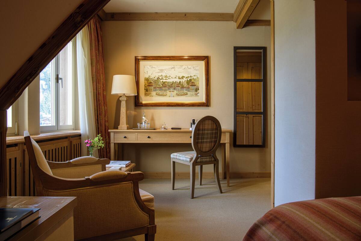 A hotel room with a wooden desk, two chairs, a lamp, a framed picture on the wall, and a window with curtains.
